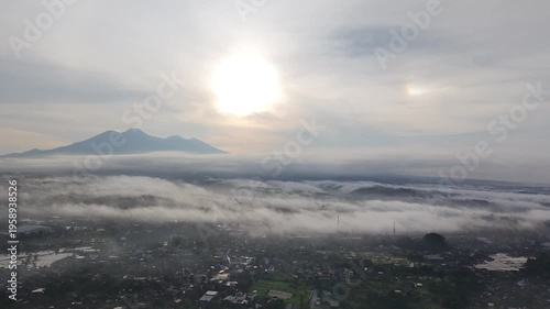 Aerial view of a mist-covered valley at sunrise, with soft clouds drifting over a town and mountains silhouetted beneath a glowing sky
