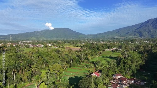 Aerial view of vibrant rice fields and a tropical village surrounded by dense greenery, with majestic mountains and a bright blue sky stretching across the horizon