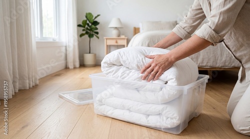 Woman hands folding white thick duvet into transparent plastic storage container on wooden floor of bright bedroom