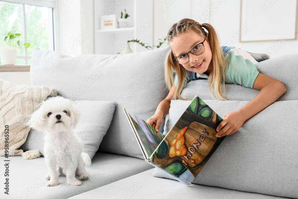 Fototapeta premium Cute little happy girl with Maltese dog reading book on sofa at home
