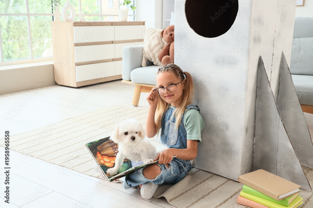Fototapeta premium Cute little happy girl with Maltese dog and books sitting on floor near cardboard space rocket at home