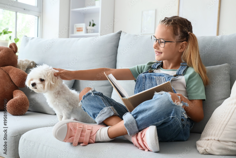 Fototapeta premium Cute little happy girl with Maltese dog reading book on sofa at home