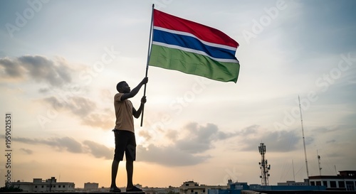 Silhouetted figure proudly raising the Gambian flag against a dramatic sunset sky, symbolizing national identity.