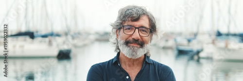 Happy male seigneur in glasses crossing his arms while standing in the port and looking at the camera on yachts background, camera is moving closer. Panoramic