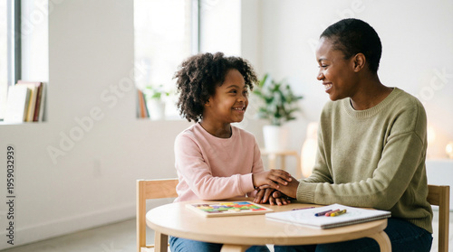 Black mother and young daughter with Down syndrome hold hands and talk at a desk, sharing a tender and educational moment at home, nurturing care, childhood development, family love.