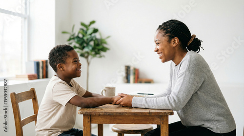An African American woman sits at a table with a young boy, holding his hands and talking during a lesson. home tutoring, communication skills, inclusive education, supportive environment.