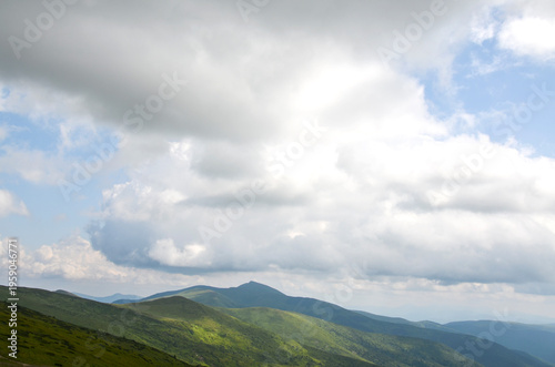 Sunlight patches illuminate a green mountain range beneath dramatic low clouds, with rolling hills and distant peaks fading into haze. Carpathian Mountains, Ukraine
