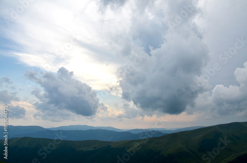 Sunlight patches illuminate a green mountain range beneath dramatic low clouds, with rolling hills and distant peaks fading into haze. Carpathian Mountains, Ukraine