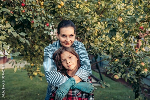 mother and daughter embrace in an orchard during harvest season. The mother is a Caucasian woman with dark hair, and the daughter is a young girl with brown hair.