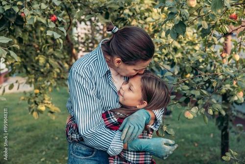mother and daughter embrace in an apple orchard during harvest. The mother is a young Caucasian woman with brown hair, and the daughter is a young girl with brown hair.