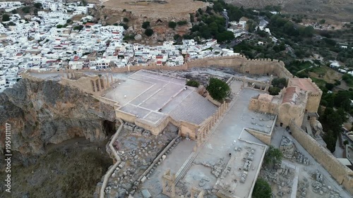 Cinematic drone orbit of the Lindos Acropolis during blue hour. The shot reveals the traditional white village and the scenic harbor at dusk. Iconic ancient landmark and Mediterranean architecture