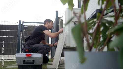 Technician servicing outdoor heat pump unit beside residential house with toolbox nearby, removing cover grille and inspecting fan. Concept of HVAC maintenance, repair and home heating and cooling
