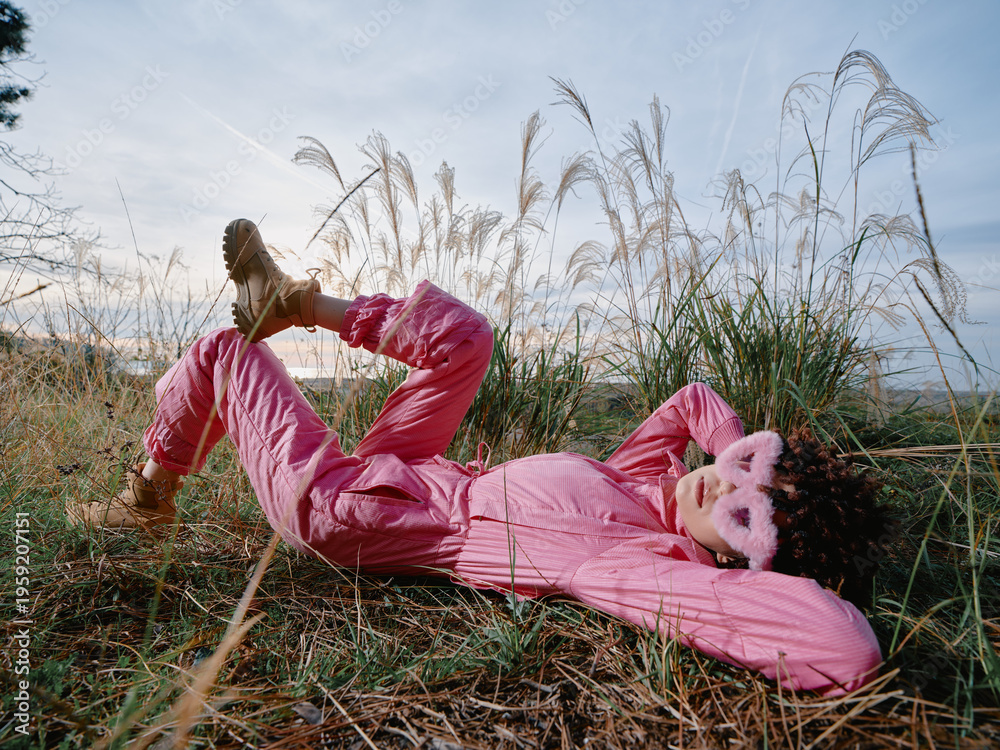 Obraz premium Outdoor fashion in a sunny meadow features a woman in a pink jumpsuit lying among tall grasses, relaxed pose, playful mood, and natural light emphasizing texture and color.