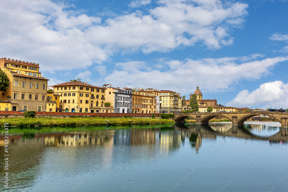 Fototapeta premium Santa Trinita Holy Trinity bridge gracefully crosses the Arno River in Florence, highlighting the beauty of medieval architecture against a backdrop of lively buildings and a blue sky.