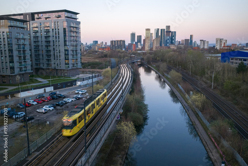 Fotografia Aerial image of a moving yellow Metrolink tram passing Pomona Wharf toward the Deansgate skyscrapers in Manchester, England, UK