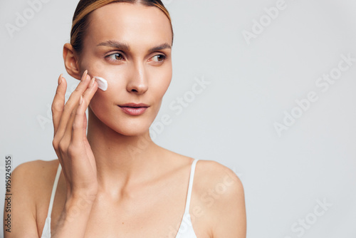 Young woman applying face cream on clean skin, skincare routine, natural beauty, soft lighting, white background, focused expression, casual wear, healthy lifestyle concept