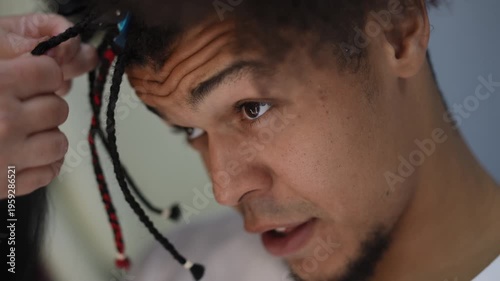 Close-up of a handsome young man getting his hair styled. A hairstylist's hands are carefully parting and braiding his afro-textured hair