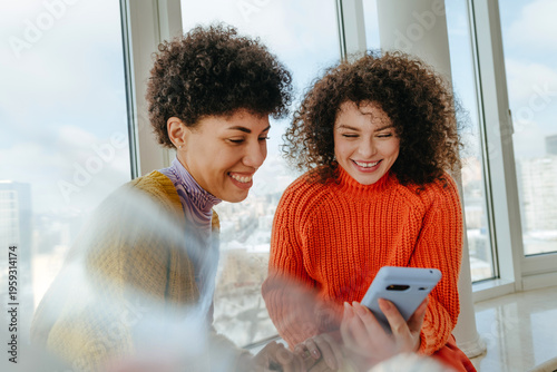 Young women with curly hair smiling and talking on windowsill indoors