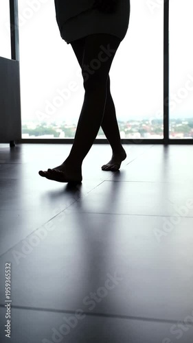 A close-up low angle shot of a woman's legs in silhouette, wrapped in a bath towel. She walks barefoot on a tiled floor towards a large floor-to-ceiling window with bright daylight and a blurred city 
