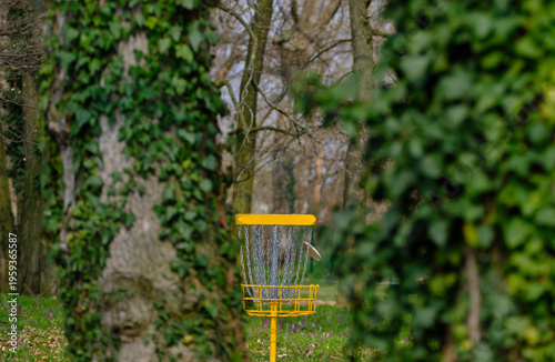 Discgolf basket with flying disc in the park between trees