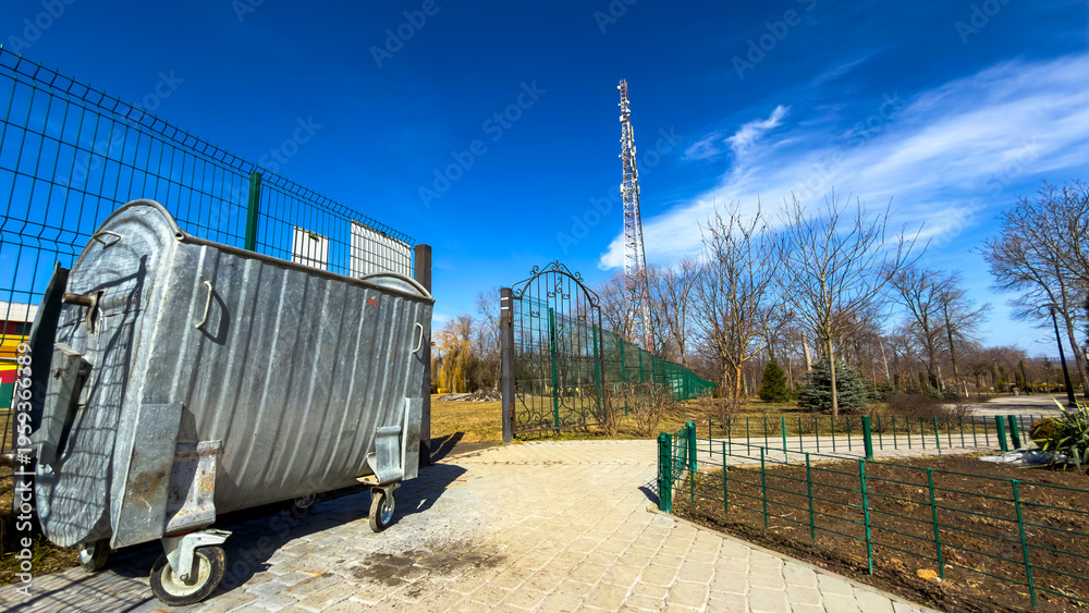 Fototapeta premium Park area with trash bin and cell tower. A trash bin stands near a park pathway with trees and a tall cell tower in the background on a sunny day.