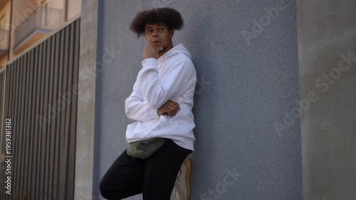 Thoughtful mixed-race man with an afro hairstyle wearing casual clothes leans against a gray wall, stroking his chin in contemplation
