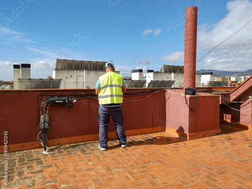 A rear view of a telecom technician in a yellow safety vest installing new fiber optic and phone lines at a junction box on a building rooftop in Barcelona.
