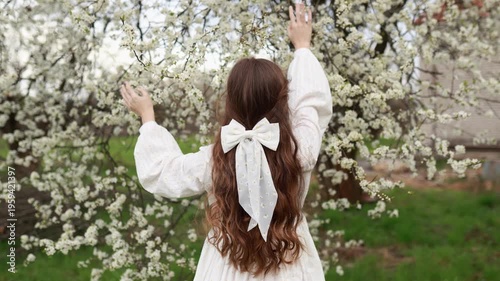 Back view of a young woman with long hair and a white bow, standing in a blooming orchard and reaching toward delicate spring blossoms. Concept of spring, beauty, femininity, and harmony with nature.