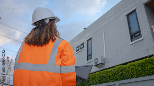 Фотография A worker in a safety helmet and orange jacket looks at a building while holding a laptop