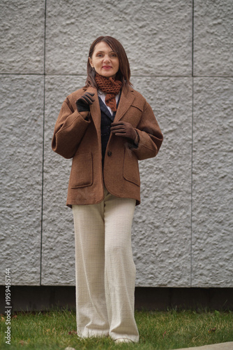A woman in autumn attire stands on grass near a modern wall. The scene represents harmony, refined taste, and the aesthetic of quiet confidence.