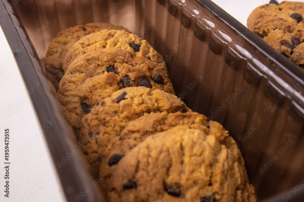 Fototapeta premium Delicious chocolate chip cookies arranged inside a dark plastic tray with a small stack on the side isolated on a white background.