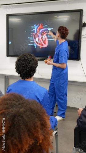 Young medical professor teaching cardiology to a diverse group of students in a classroom, pointing at a digital screen showing a diagram of the human heart and holding a physical model
