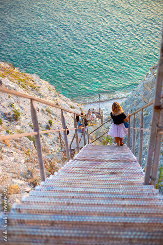 A woman in a white dress stands on a metal staircase descending to the sea. travel vacation, cliff stairs, coastal view.