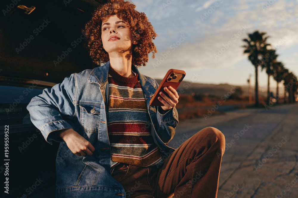 Naklejka premium A woman in a rainbow sweater and denim jacket smiles while holding a smartphone leaning against a car at sunset on a palm tree lined road. Lifestyle and technology concept.