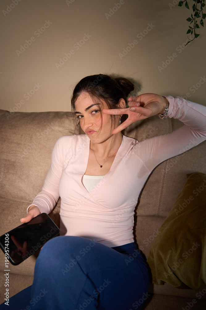 Obraz premium Young woman making peace sign holding phone on sofa