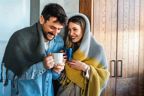 Cheerful young couple covered in blanket embracing while holding cups of tea at home