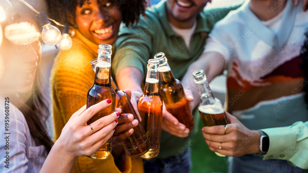 Obraz premium Happy friends enjoying night party on rooftop terrace - Group of multiracial young people drinking beer bottle at brewery pub garden - Life style concept with guys and girls hanging outdoors