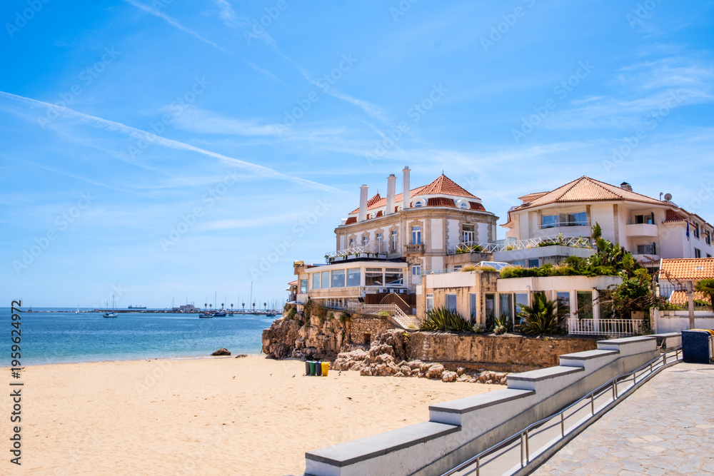 Obraz premium Beachfront villas on rocky shoreline above sandy beach Praia da Rainha in Cascais, Portugal. Wide angle view with clear sky, calm ocean, and Mediterranean architecture in bright daylight