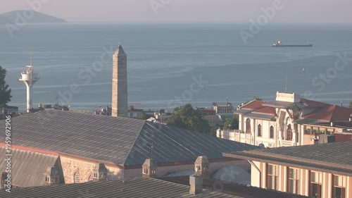 Rooftops and historic buildings overlook the calm sea with a stone tower in Istanbul under warm sunrise light. Soft colors and peaceful atmosphere create a scenic urban coastal landscape.