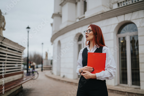 A professional woman with red hair and glasses holds a red clipboard outdoors. She stands beside a white modern building on a city street, projecting a confident, businesslike mood.