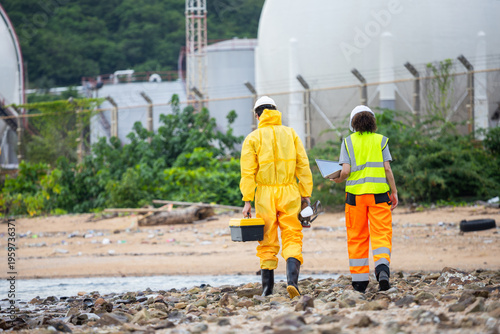Environmental Response Team in Hazmat Suit and Safety Gear Walking on Polluted Shoreline, Technicians Carrying Equipment and Laptop for Chemical Spill Assessment Near Industrial Facility
