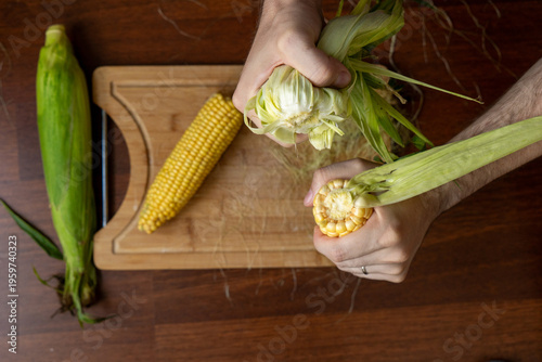 Konstfotografi Male hands holding and shucking  fresh corn on the cob on a wooden table