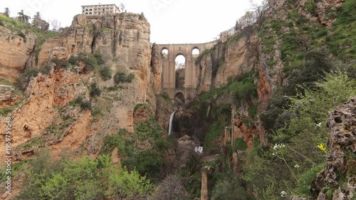 Wide view of the famous Puente Nuevo bridge in Ronda, Andalusia, spanning a deep rocky gorge with a waterfall and historical buildings on cliffs.