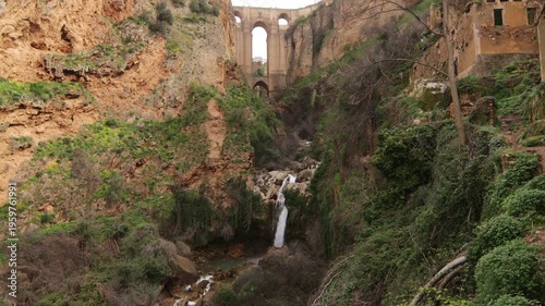 Scenic view of the iconic Puente Nuevo bridge over El Tajo gorge in Ronda, Malaga, featuring a natural waterfall and lush cliffs. Spain.