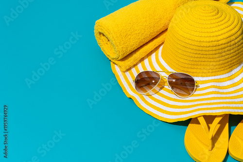 Yellow beach accessories on turquoise blue background. Bright yellow beach hat with stripes, paired with stylish sunglasses. Soft, rolled towel and vibrant yellow flip-flops. Summer vacation concept.