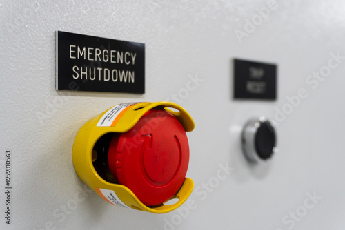 Red emergency stop button with yellow safety housing. Sign reads 'EMERGENCY SHUTDOWN'. Panel includes black push-button. Used for industrial safety or machinery control