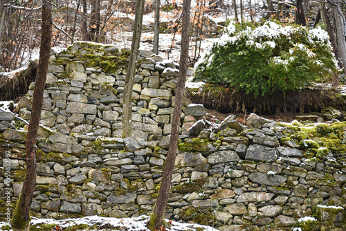 A fragment of an old stone wall made of gray stones of different sizes without visible mortar trees snow winter tourism history