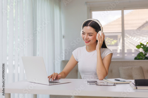 Smiling woman wearing wireless headphones, sitting at a desk and using a laptop, enjoying music or online learning