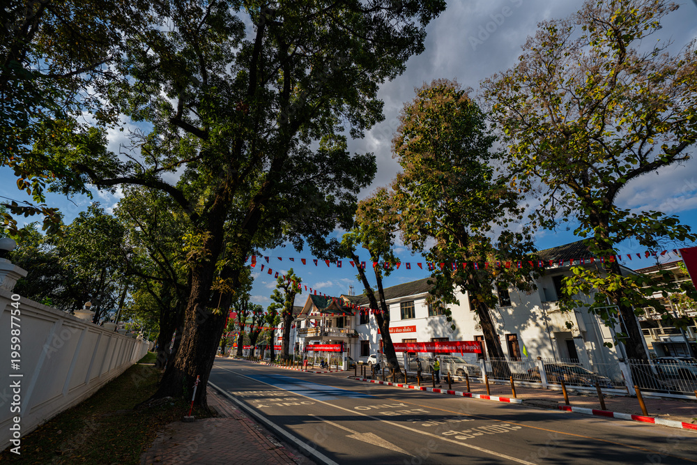 Obraz premium Vientiane Laos: 22 November 2025. A view of a sunlit urban intersection featuring light traffic in front of a prominent government building adorned with flags. The scene captures a calm city