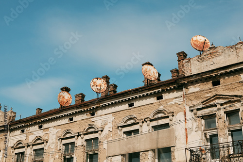 Old residential building with weathered facade and multiple satellite dishes on the roof. Urban decay, outdated infrastructure and contrast between past architecture and modern additions